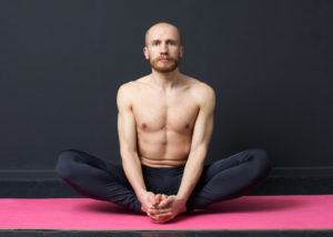 Sport and fitness. Young man with naked torso is performing groin muscle stretching - butterfly exercise on the mat, gray background
