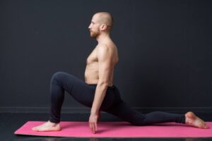 Sport and fitness. Young man with naked torso is doing front lunge on the mat, gray background
